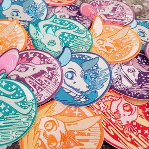 May include: A collection of embroidered patches featuring a stylized animal design. The patches showcase a variety of colors, including teal, orange, purple, and pink, with intricate details and star accents. The patches are round with a detailed border.