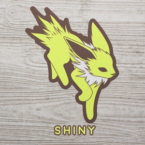 May include: A yellow and brown shiny Eevee Pokemon character illustration with the word "SHINY" printed below.