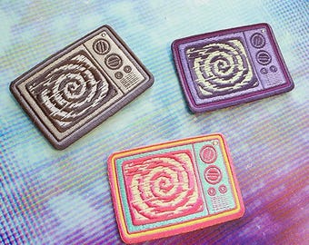 May include: Three rectangular embroidered patches shaped like vintage television sets. Each patch features a central spiral pattern on the screen and two circular knobs on the right side. The patches are colored in brown, purple, and pink.