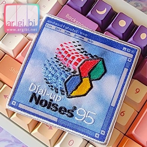 May include: Embroidered patch featuring a retro Windows 95 design with the text "Dial-up Noises '95". The patch has a blue background with a colorful logo and is surrounded by a blue border. The patch is placed on a keyboard.