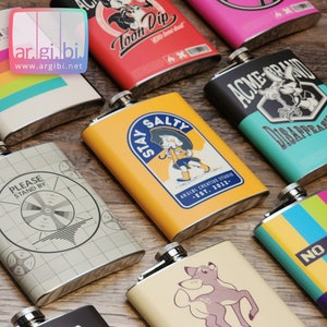 Puede incluir: Una colección de matraces de acero inoxidable con varios diseños coloridos. Los matraces presentan diferentes ilustraciones y textos, incluyendo "Stay Salty", "Please Stand By", "Toon Dip", "Acme-Brand" y "No".