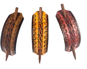May include: Three curved leather hair slides in tan, yellow, and reddish-brown, each featuring a central row of embossed geometric patterns and a dark brown wooden stick pin inserted through the leather to secure the hair accessory in place.