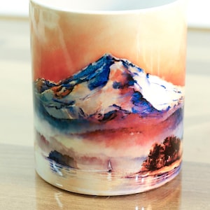 May include: A white ceramic mug with a watercolor painting of a mountain range at sunset. The mountain peaks are covered in snow and the sky is a vibrant orange and pink.