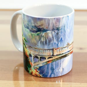 May include: White ceramic mug with a watercolor design of a bridge over a waterfall. The bridge is made of stone and has arches. The waterfall is blue and white and has a cloudy sky above it.