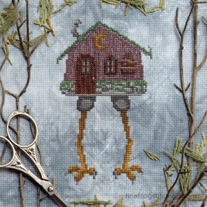 May include: A cross-stitch pattern of a pink house with a green roof and a crescent moon on the front. The house is on two long, yellow legs with brown feet. The background is a light blue with brown branches.