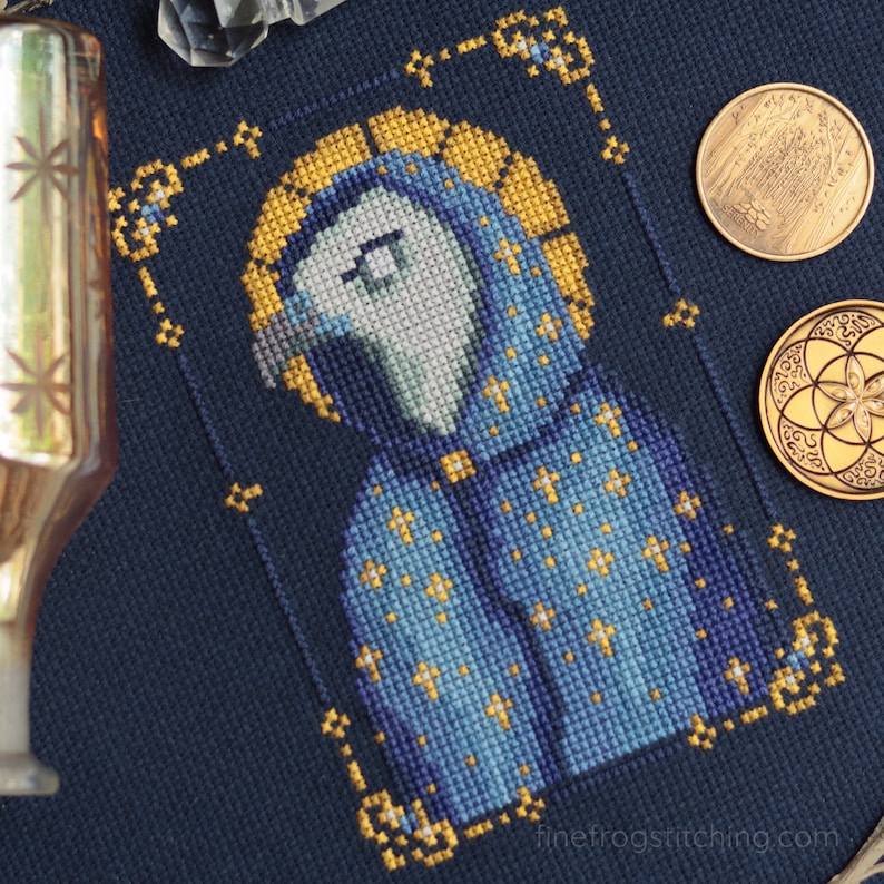 Pode incluir: Uma obra de arte em ponto cruz que retrata uma figura em uma t&uacute;nica azul com detalhes dourados e uma aur&eacute;ola. A figura &eacute; cercada por uma borda dourada e pequenas cruzes douradas. O fundo &eacute; um tecido azul escuro. Duas moedas de ouro s&atilde;o vis&iacute;veis.