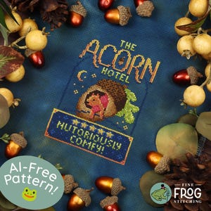 May include: A cross-stitch pattern on dark blue fabric featuring an acorn-shaped building with the text THE ACORN HOTEL and NUTORIOUSLY COMFY, surrounded by a border of scattered acorns, pinecones, and small yellow berries.