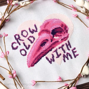 May include: Cross stitch pattern with a pink and purple crow skull in a heart shape with the text "Grow Old With Me".