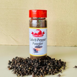 K&ouml;nnte beinhalten: Ein durchsichtiges Kunststoff-Gew&uuml;rzglas mit rotem Deckel, gef&uuml;llt mit dunkelbraunem Kubebenpfeffer-Pulver. Das wei&szlig;e Etikett tr&auml;gt den Text Planet Spices, Cubeb Pepper Powder und Net Wt.: 100 g. Ein Haufen ganzer getrockneter Kubebenpfefferk&ouml;rner liegt um die Basis.