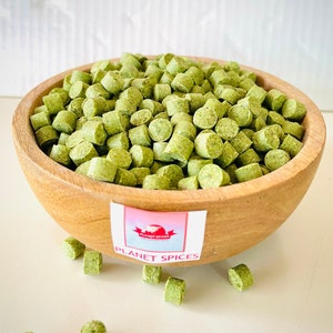 Puede incluir: Un cuenco de madera lleno de pequeños trozos de comida cilíndricos, secos y verdes. El cuenco está sobre una superficie blanca. El texto "PLANET SPICES" está en una etiqueta debajo del cuenco.