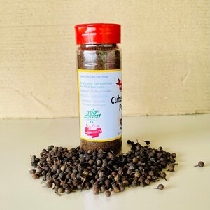 K&ouml;nnte beinhalten: Ein durchsichtiges Kunststoff-Gew&uuml;rzglas mit rotem Deckel, gef&uuml;llt mit dunkelbraunem Kubebenpfeffer-Pulver. Das wei&szlig;e Etikett tr&auml;gt den Text "Cubeb Pepper Powder" und "100% Natural Product". Ein Haufen ganzer, dunkler Kubebenpfefferk&ouml;rner liegt davor.