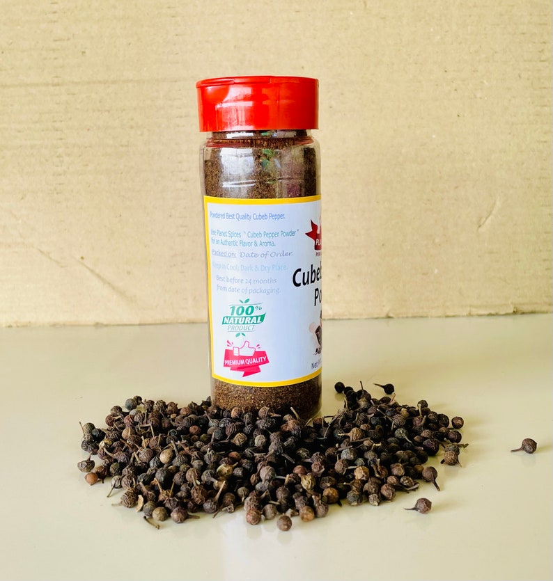 K&ouml;nnte beinhalten: Ein durchsichtiges Kunststoff-Gew&uuml;rzglas mit rotem Deckel, gef&uuml;llt mit dunkelbraunem Kubebenpfeffer-Pulver. Das wei&szlig;e Etikett tr&auml;gt den Text "Cubeb Pepper Powder" und "100% Natural Product". Ein Haufen ganzer, dunkler Kubebenpfefferk&ouml;rner liegt davor.