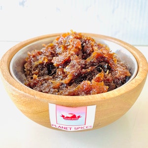 May include: A wooden bowl with a silver interior contains a portion of a dark brown, glistening food item, possibly a chutney or relish. The bowl has a label that reads "PLANET SPICES" with a red bird logo.
