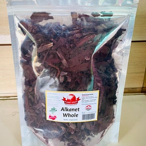 May include: A clear, resealable plastic pouch filled with dark brown, dried Alkanet root pieces. A white label on the front features the text Planet Spices, Alkanet Whole, and Net Wt.: 100 g (3.53 oz), along with a barcode and contact details.