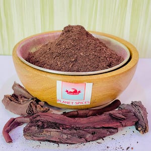 May include: A wooden bowl filled with a dark brown powder, likely a spice blend. The bowl is sitting on a white surface with pieces of dark brown bark nearby. The label on the bowl reads "Planet Spices".