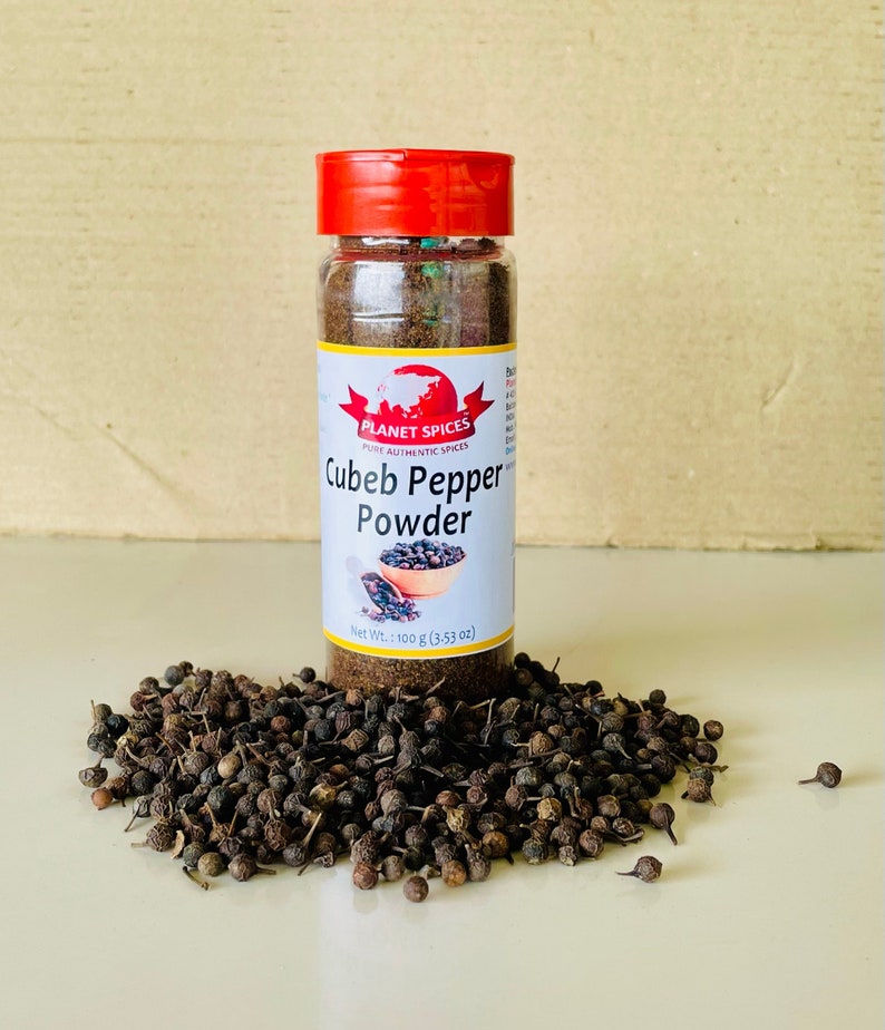 K&ouml;nnte beinhalten: Ein durchsichtiges Kunststoff-Gew&uuml;rzglas mit rotem Deckel, gef&uuml;llt mit dunkelbraunem Kubebenpfeffer-Pulver. Das wei&szlig;e Etikett tr&auml;gt den Text Planet Spices, Cubeb Pepper Powder und Net Wt.: 100 g. Ein Haufen ganzer getrockneter Kubebenpfefferk&ouml;rner liegt um die Basis.