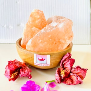 May include: A wooden bowl filled with large, translucent pink Himalayan salt chunks. A white label on the bowl features a logo and the text PLANET SPICES. Two dried, deep red hibiscus flowers and several loose purple petals are arranged nearby.