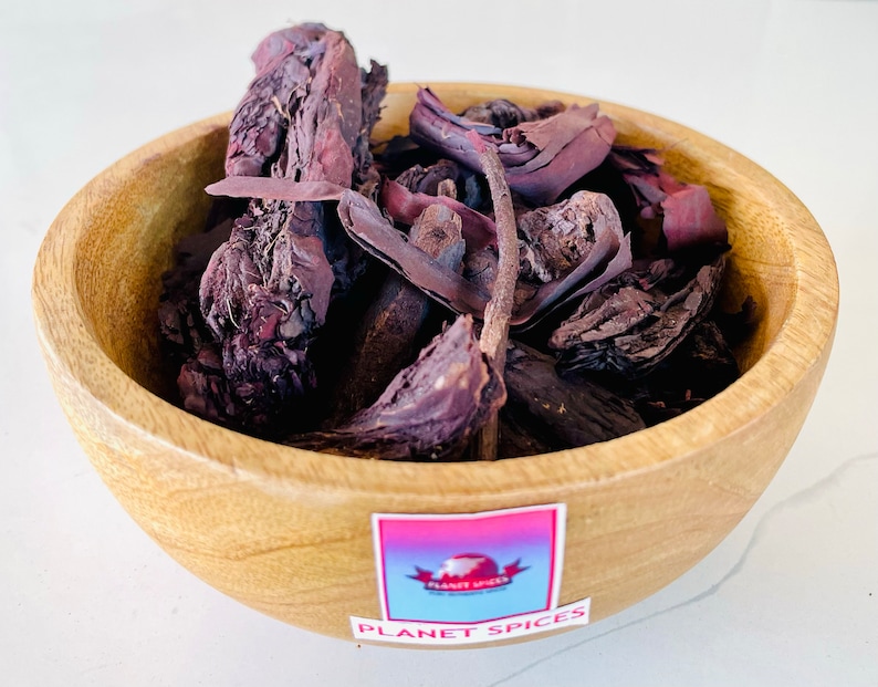 May include: A small, round, light-brown wooden bowl filled with dark purple, dried, woody bark pieces. A square sticker on the front of the bowl features a gradient background, a red logo, and the text PLANET SPICES in white capital letters.