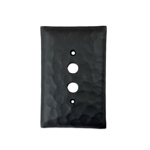 May include: A black, rectangular light switch cover with a hammered texture. It has two round holes for the light switches and one smaller hole above the bottom switch. The cover is made of a dark, matte material.