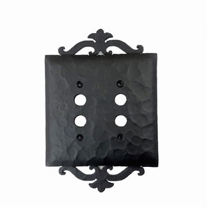 May include: Black, textured double light switch cover with decorative flourishes. The square cover has a hammered appearance and features four circular openings for the light switches. The decorative elements are at the top and bottom of the cover.