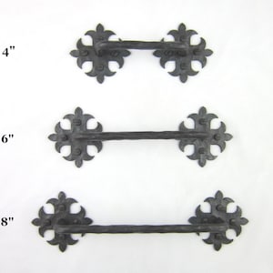 May include: Three wrought iron handles in varying sizes: 4-inch, 6-inch, and 8-inch. Each handle features a central bar with decorative, fleur-de-lis style end pieces. The metal has a dark, textured finish, suggesting a rustic or antique aesthetic.