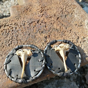 Puede incluir: Un par de pendientes de cuero negro con un dije de hueso. Los pendientes están hechos de cámaras de aire recicladas y tienen un aro de metal plateado.