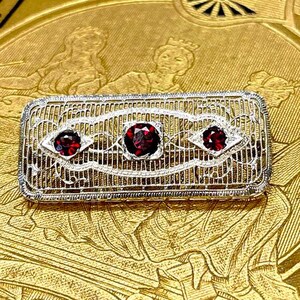 May include: A rectangular antique brooch with three red gemstones set in a filigree design. The brooch is silver-toned with intricate detailing. The background features gold-toned text that reads "USEUM OF ANTIQ".