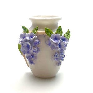 May include: A small, off-white ceramic vase with a rounded body and a flared rim. The vase is adorned with clusters of lavender-colored flowers and green leaves, adding a touch of floral elegance. The vase is a decorative item.