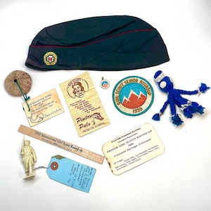 May include: A collection of vintage Girl Scout memorabilia, including a dark green hat with a red stripe, patches, wooden tags, and a small figurine. The items are arranged on a white surface, showcasing a nostalgic theme.