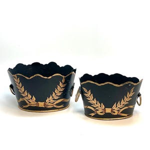 May include: Two oval-shaped black metal planters with scalloped top edges and gold-painted laurel leaf and ribbon motifs. Each planter features small gold-toned metal ring handles on the sides, providing a decorative accent to the dark finish.