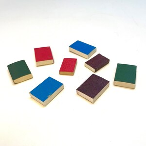 Op de afbeelding: Acht miniatuurboekjes met verschillende gekleurde omslagen. De omslagen zijn rood, blauw, groen, paars en bruin. De boekjes zijn in een cirkel op een wit oppervlak gerangschikt.