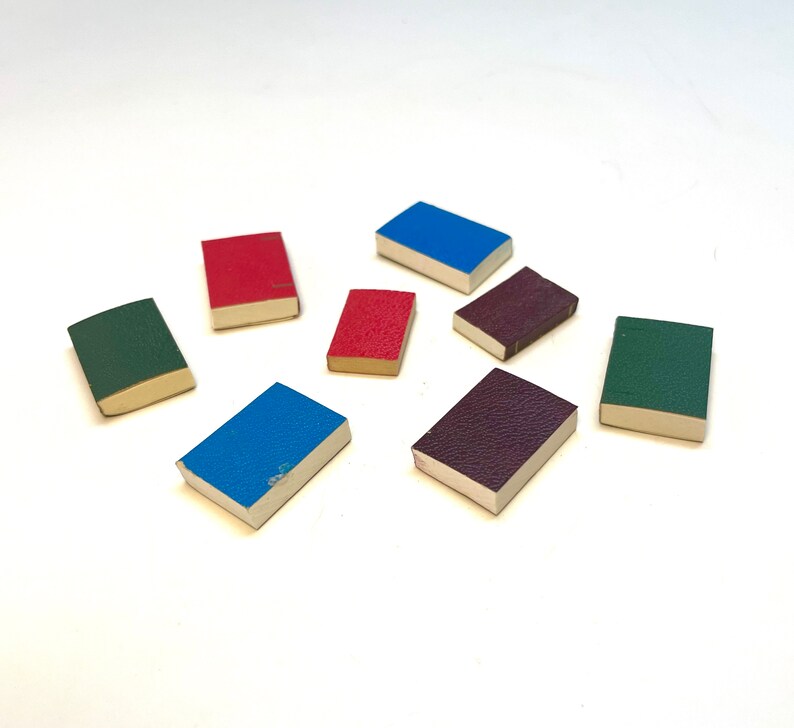 Op de afbeelding: Acht miniatuurboekjes met verschillende gekleurde omslagen. De omslagen zijn rood, blauw, groen, paars en bruin. De boekjes zijn in een cirkel op een wit oppervlak gerangschikt.