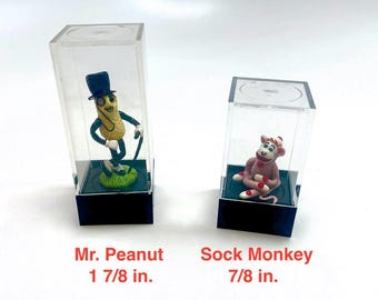 Op de afbeelding: Twee nieuwe beeldjes, Mr. Peanut en een Sok-aap, tentoongesteld in doorzichtige plastic koffers met zwarte bases. Mr. Peanut, 4,76 cm hoog, draagt een hoge hoed en een monocle. De Sok-aap, 2,22 cm hoog, is roze en rood.