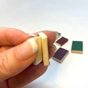 Op de afbeelding: Een hand die twee kleine, witte, rechthoekige blokken vasthoudt. Er liggen drie andere kleine, rechthoekige blokken op de tafel, een blauwe, een groene en een paarse.