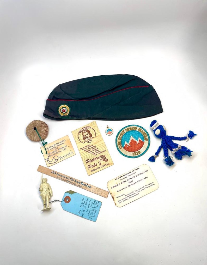 May include: A collection of vintage Girl Scout memorabilia, including a dark green hat with a red stripe, patches, wooden tags, and a small figurine. The items are arranged on a white surface, showcasing a nostalgic theme.