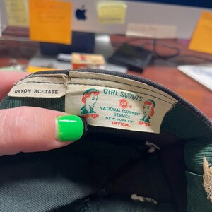 May include: Close-up of a vintage Girl Scouts cap interior. The label reads "Girl Scouts National Equipment Service New York City Official." The cap is dark green with a green and white label. The label features two illustrations of Girl Scouts.