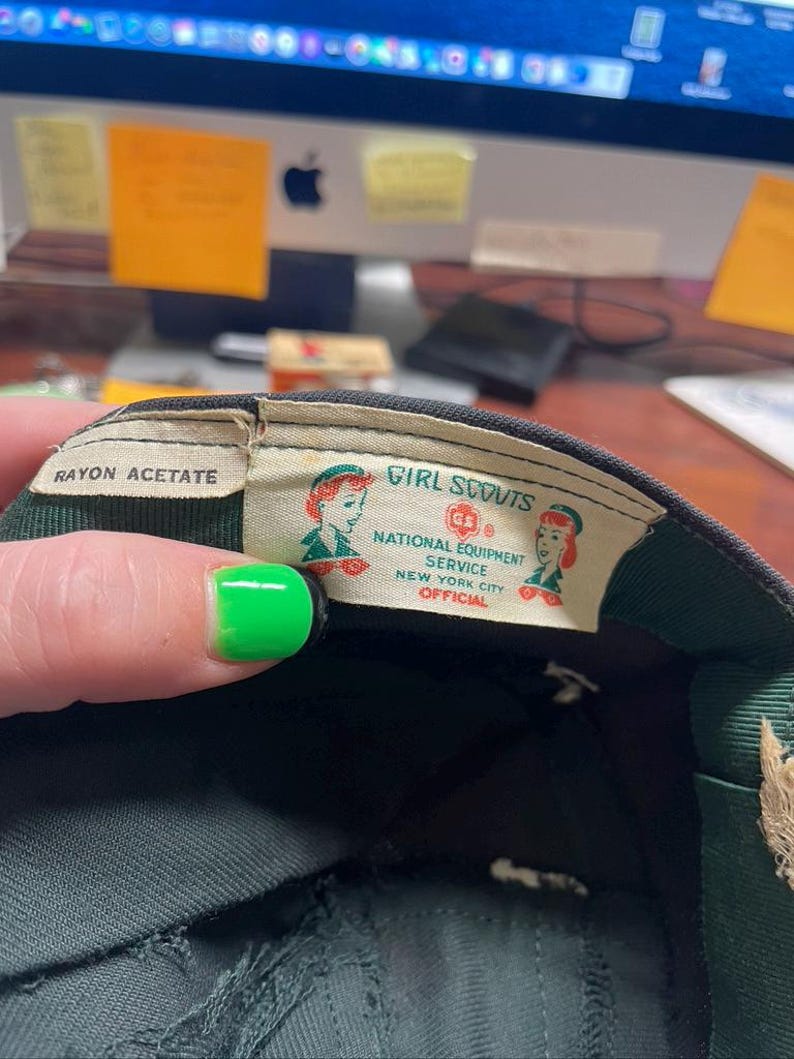 May include: Close-up of a vintage Girl Scouts cap interior. The label reads "Girl Scouts National Equipment Service New York City Official." The cap is dark green with a green and white label. The label features two illustrations of Girl Scouts.