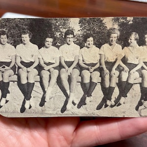 May include: A vintage black and white photograph of seven people seated on a bench. They are wearing matching light-colored shirts, dark shorts, and knee-high socks. The image has a slightly aged appearance.