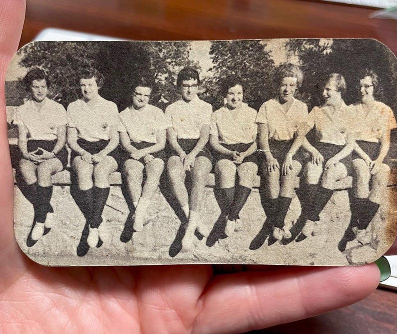 May include: A vintage black and white photograph of seven people seated on a bench. They are wearing matching light-colored shirts, dark shorts, and knee-high socks. The image has a slightly aged appearance.