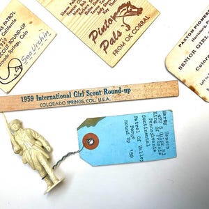 May include: A collection of vintage Girl Scout memorabilia, including tags, a wooden ruler with text, and a small plastic figurine. The tags feature text and illustrations, with the words "Girl Scout Round-up" and "Pinto Pals".