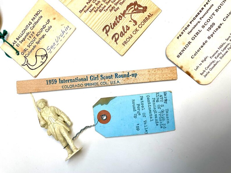 May include: A collection of vintage Girl Scout memorabilia, including tags, a wooden ruler with text, and a small plastic figurine. The tags feature text and illustrations, with the words "Girl Scout Round-up" and "Pinto Pals".