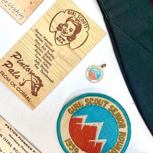 May include: A collection of vintage Girl Scout memorabilia, including patches, pins, and wooden name tags. The items feature the Girl Scout logo and text, with a focus on the 1959 Girl Scout Senior Roundup.