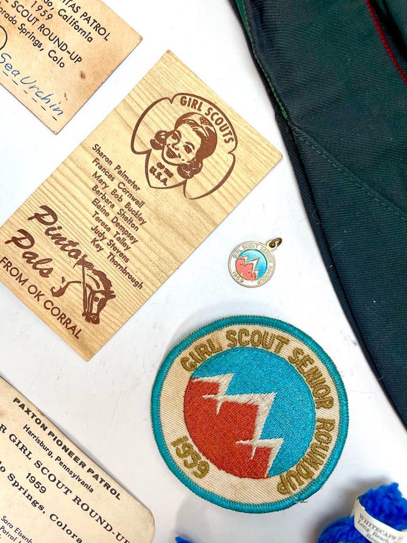 May include: A collection of vintage Girl Scout memorabilia, including patches, pins, and wooden name tags. The items feature the Girl Scout logo and text, with a focus on the 1959 Girl Scout Senior Roundup.