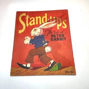 May include: A vintage children's book cover featuring a white rabbit wearing a blue jacket and brown pants. The title is "Stand-Ups The tale of Peter Rabbit" with the author's name "Sidney Sage" at the bottom. The cover is red with a wood floor background.