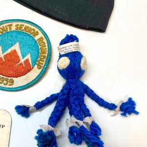 May include: A blue yarn doll with white accents and a white label that reads "WHITECAPS PATH Long Beach, Calif. Roundup 1959." A Scout Senior Roundup patch and a dark green fabric are also visible.