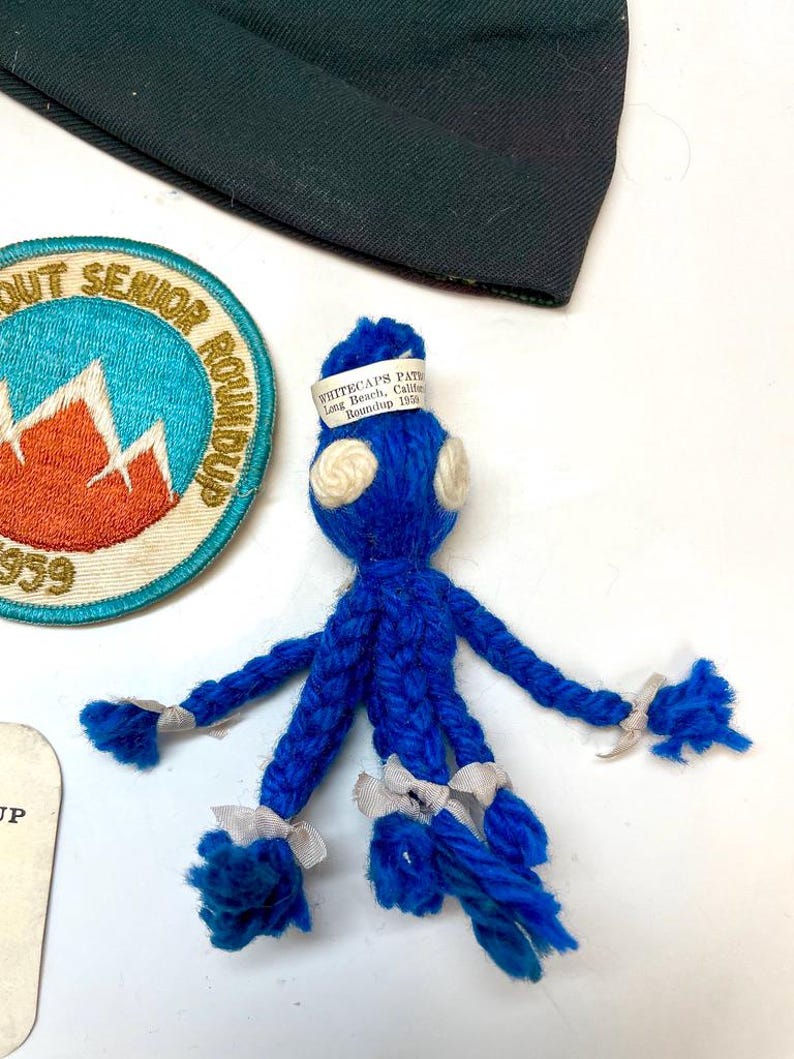May include: A blue yarn doll with white accents and a white label that reads "WHITECAPS PATH Long Beach, Calif. Roundup 1959." A Scout Senior Roundup patch and a dark green fabric are also visible.