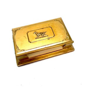 May include: A rectangular gold-toned metal memo pad holder featuring an engraved illustration of a dog on the lid. The lid has decorative corner accents and a central rectangular border surrounding the dog graphic. The base holds a stack of paper.
