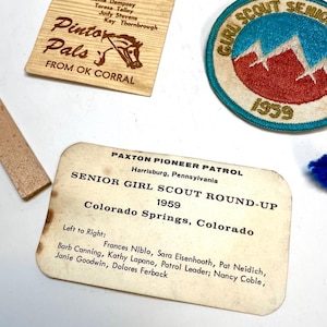 May include: A collection of vintage Girl Scout memorabilia, including a patch with a mountain design and the text "Girl Scout Senior 1959." Also present are wooden plaques with text and a pin with a mountain design.