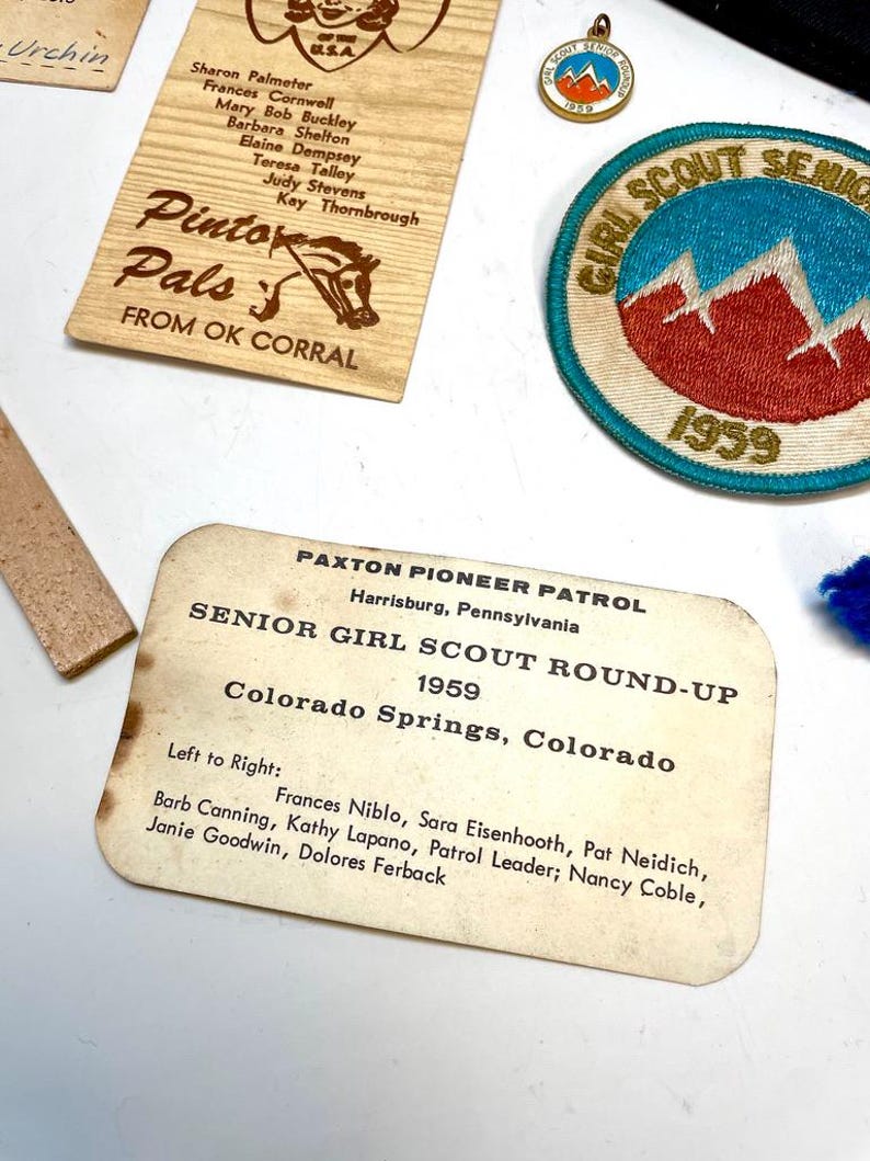 May include: A collection of vintage Girl Scout memorabilia, including a patch with a mountain design and the text "Girl Scout Senior 1959." Also present are wooden plaques with text and a pin with a mountain design.