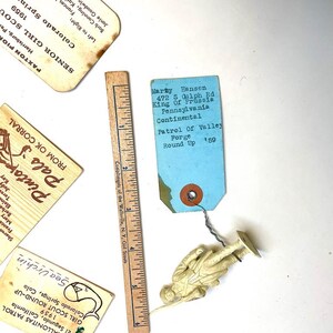 May include: A collection of vintage Girl Scout memorabilia, including tags, a wooden ruler, and a small plastic toy soldier. The tags feature handwritten text and the words "Sea Urchin" and "Round Up". The ruler measures up to 6 inches.
